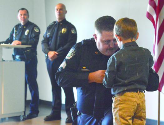 KYLE CRAVEN kneels as his son Matthew pins a sergeant’s badge on him. Capt. Robert Warner and Chief Greg Dull, in the background, lead the July 27 promotion ceremony, advancing four officers from the rank of corporal to sergeant.
