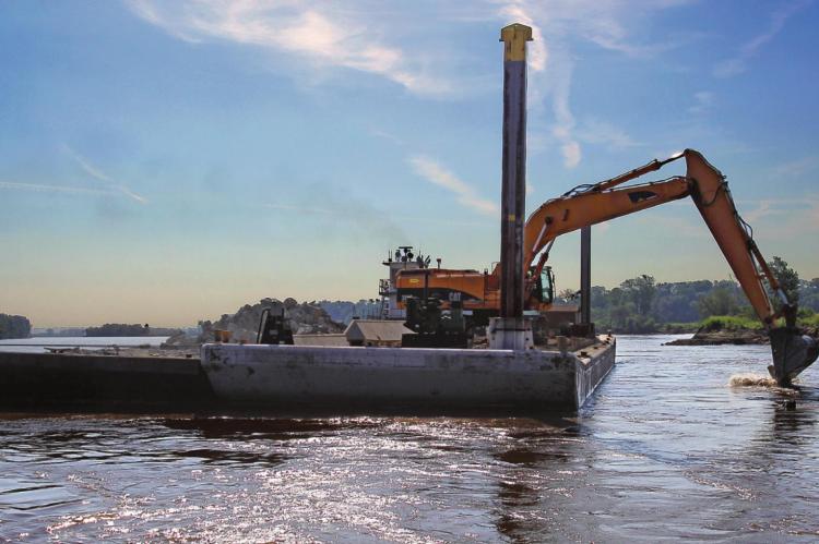 ALONG THE SOUTH bank of the Missouri River, an excavator moves rock from off the barge and into the river to rebuild a flood-damaged dike that supports river commerce.