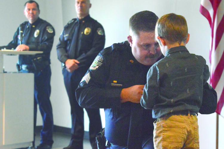KYLE CRAVEN kneels as his son Matthew pins a sergeant’s badge on him. Capt. Robert Warner and Chief Greg Dull, in the background, lead the July 27 promotion ceremony, advancing four officers from the rank of corporal to sergeant.